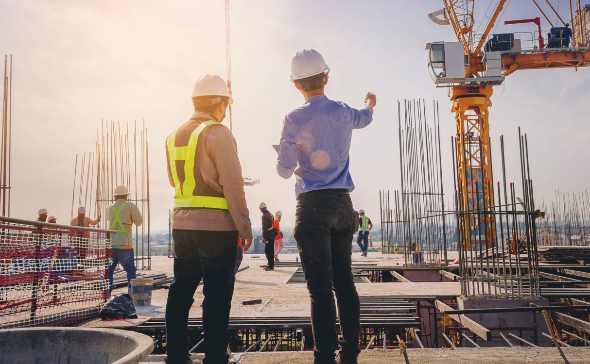 Two construction professionals wearing safety gear and helmets discussing plans on an active building site with a crane in the background.